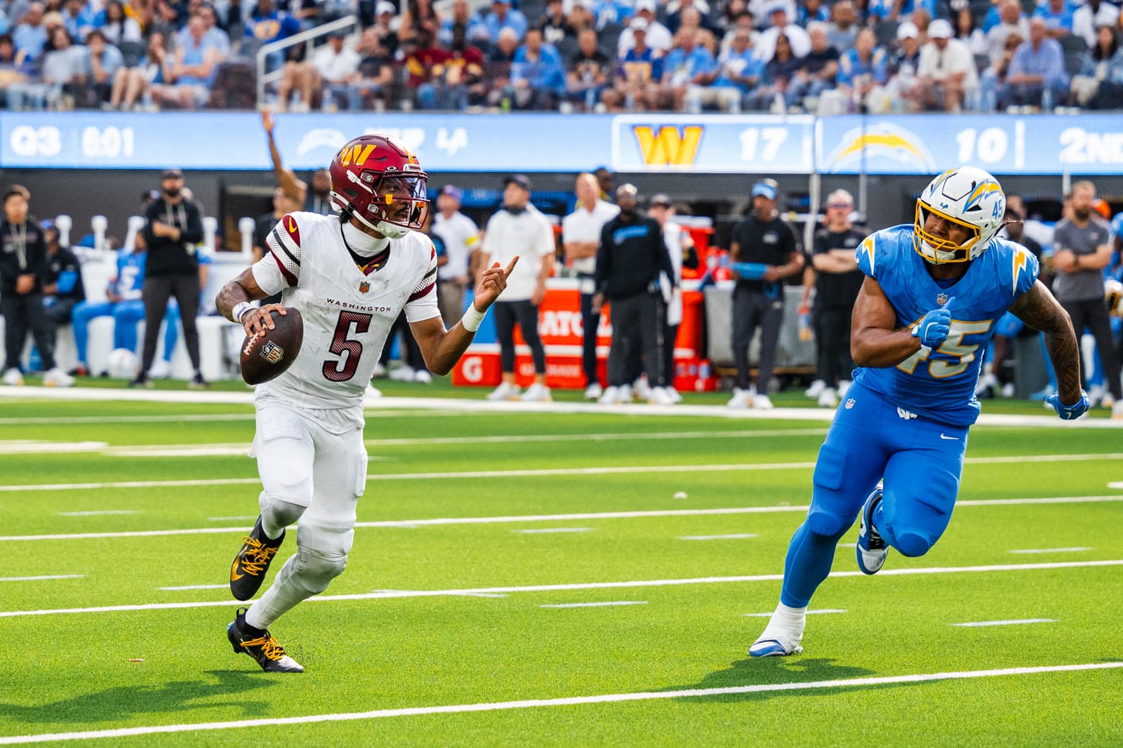 Jayden Daniels, No. 5 in a white Commanders uniform, rolls out holding the ball and pointing downfield as a Chargers pass rusher closes in.