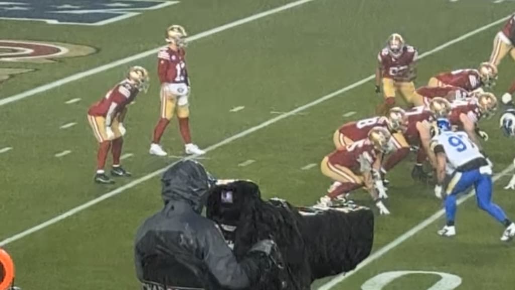 Brock Purdy, No. 13 in a red 49ers uniform, stands in the shotgun as his offensive line crouches at the line against the Rams in the rain.
