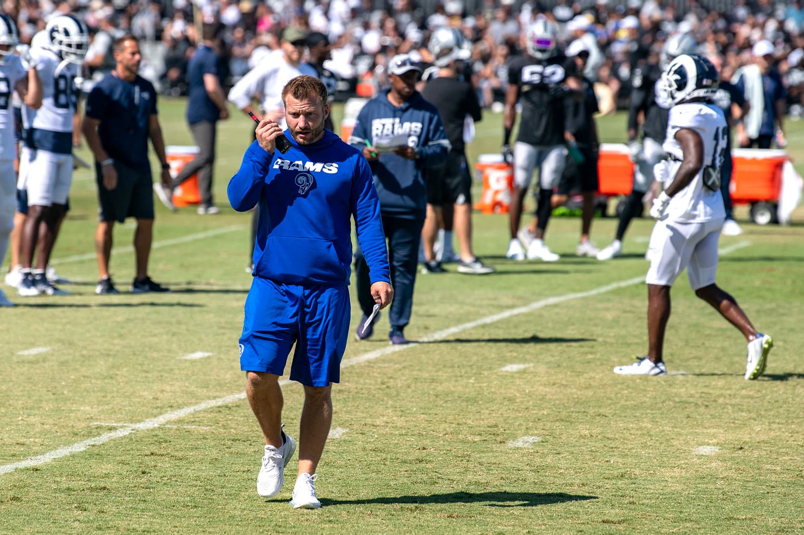 Sean McVay in a blue Rams hoodie and shorts talks into a radio while walking across a practice field, with Rams and Raiders players behind him.
